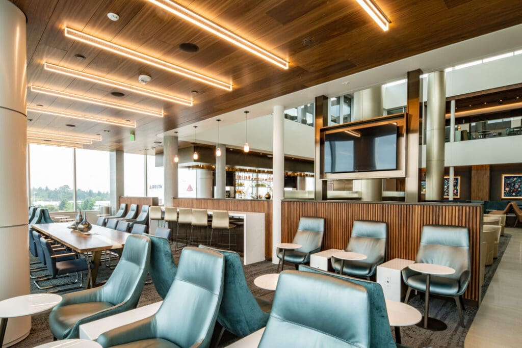 The image shows a modern airport lounge with sleek, contemporary design elements. There are comfortable blue chairs with small round tables in front of them, arranged in a seating area. A long wooden table with chairs is visible on the left side. The ceiling features wooden panels with linear lighting. A bar area with high stools and hanging pendant lights is in the background. A large screen is mounted on a wooden partition. Large windows provide natural light and a view of the outside.