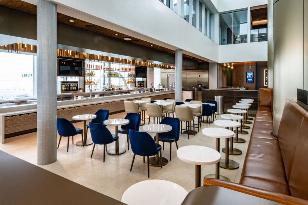 The image shows a modern, upscale airport lounge with a bar area. There are rows of tables with blue and beige chairs, and a long brown leather bench with small round tables. The bar is well-stocked with bottles and has a sleek, contemporary design. The space is well-lit with natural light coming through large windows.
