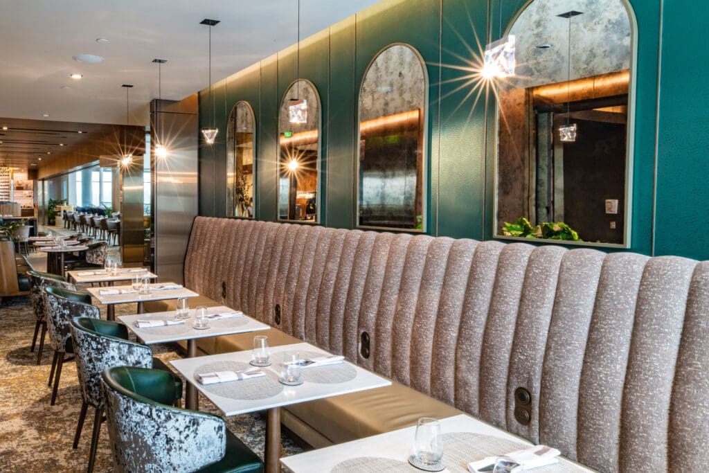 The image shows a modern, upscale restaurant interior. There is a long, upholstered bench with a textured fabric along one wall, accompanied by several tables set with placemats, napkins, and glassware. The wall features large, arched mirrors and pendant lights hanging from the ceiling. The decor includes green and brown tones, with a patterned carpet on the floor. The space appears elegant and well-lit, with additional seating visible in the background.