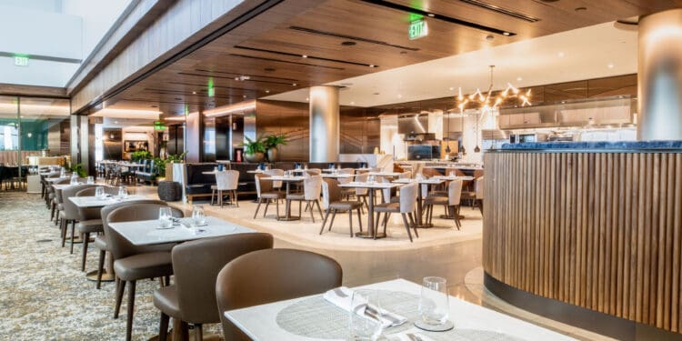 The image shows a modern, upscale restaurant interior. The space features neatly arranged tables with white tablecloths, set with glassware and napkins. The chairs are upholstered in a light brown color. The restaurant has a warm ambiance with wooden paneling on the ceiling and walls, and a patterned carpet on the floor. There are decorative plants and contemporary lighting fixtures, including a chandelier. The open kitchen area is visible in the background.
