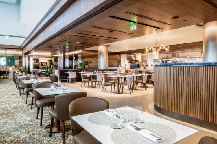 The image shows a modern, upscale restaurant interior. The space features neatly arranged tables with white tablecloths, set with glassware and napkins. The chairs are upholstered in a light brown color. The restaurant has a warm ambiance with wooden paneling on the ceiling and walls, and a patterned carpet on the floor. There are decorative plants and contemporary lighting fixtures, including a chandelier. The open kitchen area is visible in the background.