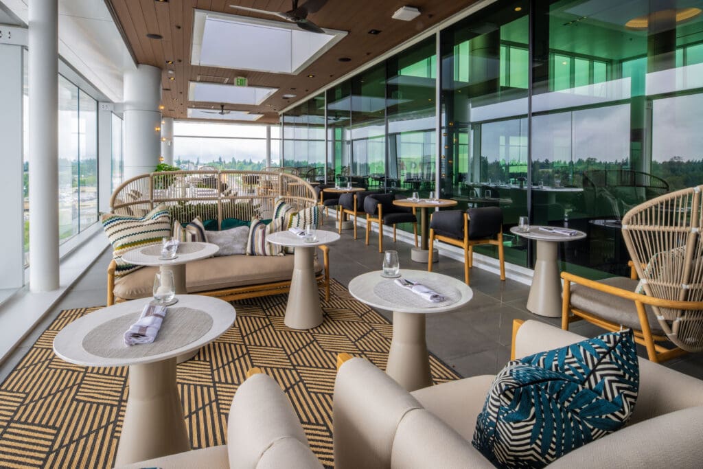 The image shows a modern, stylish indoor lounge area with large windows offering a view of the outdoors. The space features a variety of seating options, including cushioned chairs and sofas with patterned pillows. There are small round tables set with glasses and napkins. The decor includes a patterned rug and a wooden ceiling with recessed lighting. The overall atmosphere is bright and inviting.