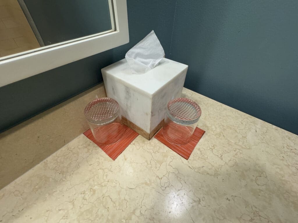 Hotel Indigo San Diego Gaslamp Quarter Studio Suite. The image shows a bathroom countertop with a marble tissue box in the center. On either side of the tissue box, there are two clear glasses placed on red coasters. A mirror is partially visible on the left side of the image, and the wall is painted a dark teal color.
