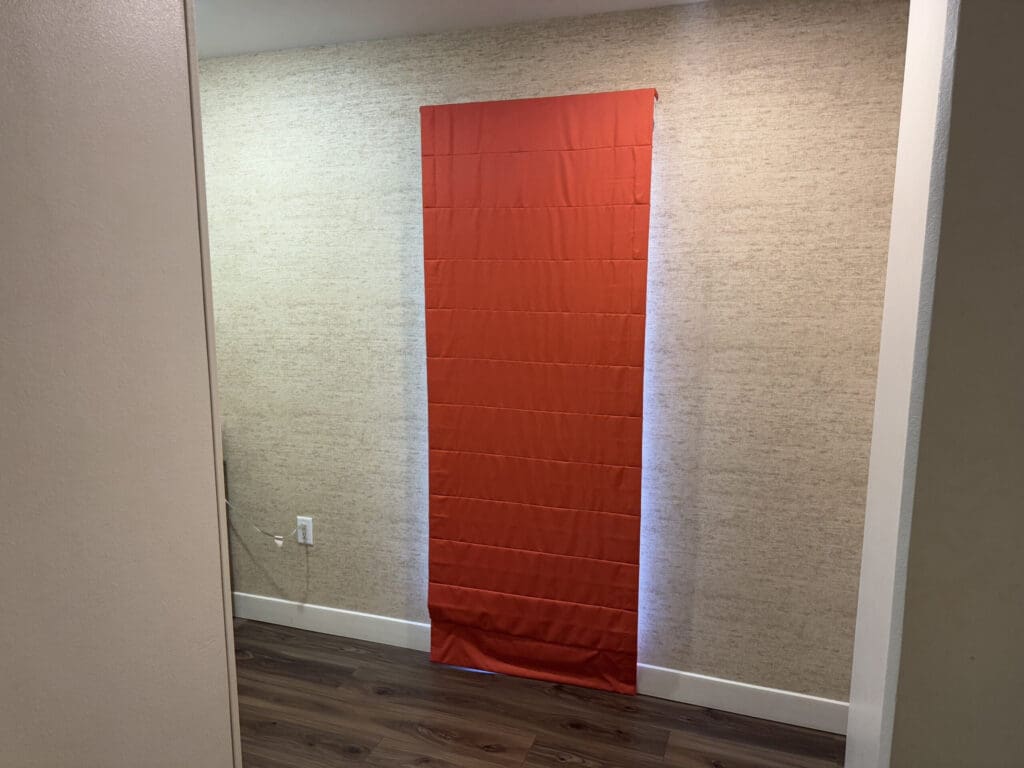 Hotel Indigo San Diego Gaslamp Quarter Studio Suite: A red curtain is hanging in front of a window on a beige textured wall. The floor is made of dark wood, and there is a white electrical outlet visible on the wall. The image is taken from an angle showing part of an adjacent wall on the left.