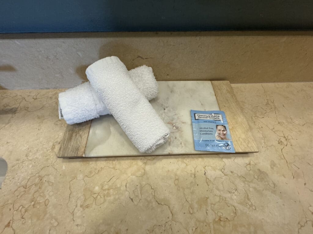 Hotel Indigo San Diego Gaslamp Quarter Studio Suite. The image shows two rolled white towels placed on a marble surface with wooden edges. Next to the towels is a blue packet labeled "Cleansing & Makeup Remover Towelettes." The surface is on a beige marble countertop.