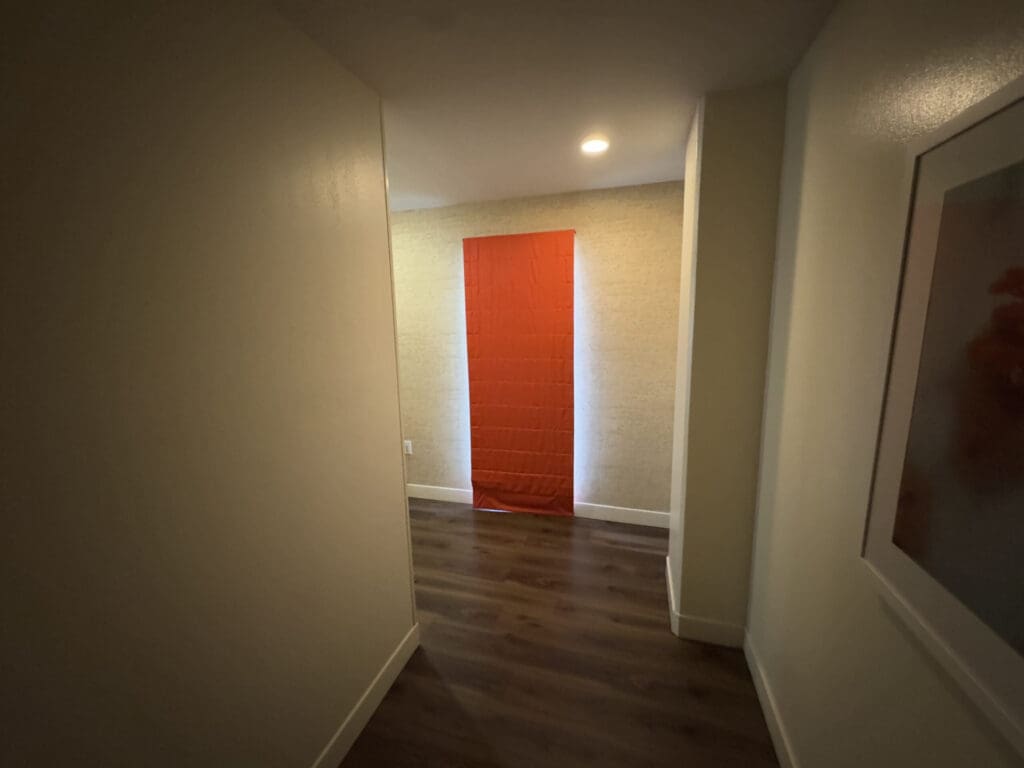 A hallway with wooden flooring and light-colored walls. At the end of the hallway, there is a bright red panel or curtain against a textured wall. A framed picture is partially visible on the right wall. The area is lit by a ceiling light.