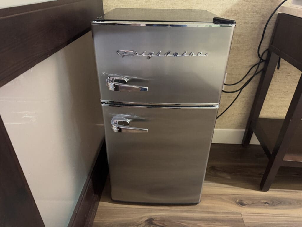 Hotel Indigo San Diego Gaslamp Quarter Studio Suite. A small, silver Frigidaire mini fridge with two doors is placed in a corner on a wooden floor. The fridge has chrome handles and is positioned between a wall and a piece of furniture. Electrical cords are visible behind it.