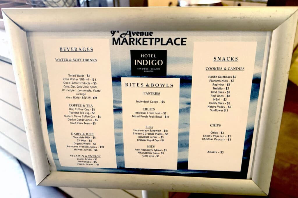 The image shows a framed menu from the "9th Avenue Marketplace" at Hotel Indigo in San Diego. The menu is divided into three sections: "Beverages," "Bites & Bowls," and "Snacks." - **Beverages** include water and soft drinks, coffee and tea, dairy and juice, and vitamin and energy drinks, with prices ranging from $5 to $15. - **Bites & Bowls** feature pastries, fruits, and various bites like sandwiches and cheese plates, with prices from $2 to $10. It also lists some medications. - **Snacks** are categorized into cookies and candies, and chips, with prices from $3 to $8.