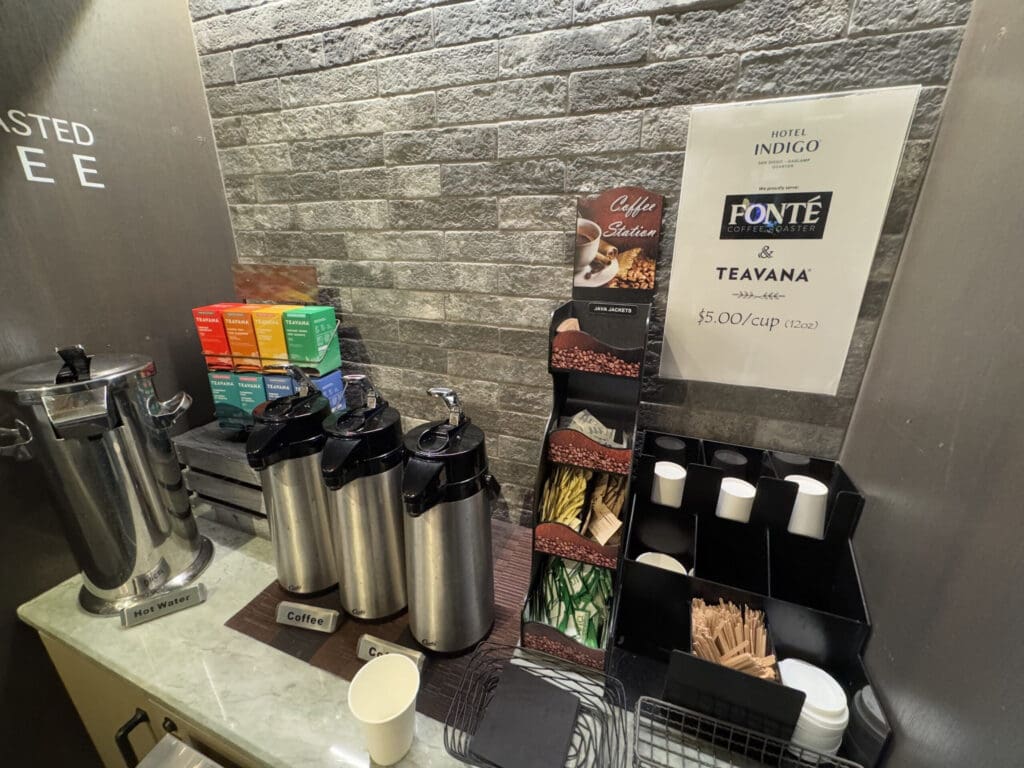 The image shows a coffee and tea station set against a brick wall. There are three stainless steel dispensers labeled "Coffee" and one labeled "Hot Water." A variety of Teavana tea boxes are stacked on the left. A sign on the wall reads "Hotel Indigo" and mentions Fonte Coffee Roaster and Teavana, with a price of $5.00 per cup (12oz). There are also compartments holding cups, lids, stirrers, and sweeteners.