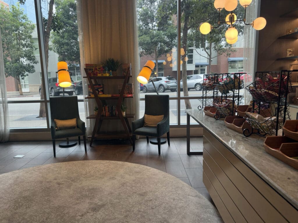 The image shows a cozy indoor seating area with two armchairs and a small bookshelf between them, placed in front of large windows. The bookshelf contains various books and small plants. Two orange lamps are positioned near the chairs. To the right, there is a counter with wire racks holding snacks. A modern chandelier with round bulbs hangs from the ceiling. Outside the windows, trees and parked cars are visible on the street.