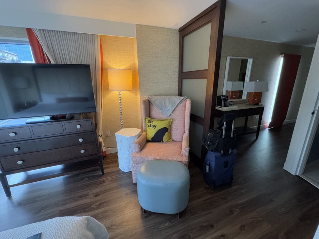 Hotel Indigo San Diego Gaslamp Quarter Studio Suite. The image shows a hotel room interior with a modern design. There is a large flat-screen TV on a dark wooden dresser with drawers. Next to it is a tall floor lamp with a warm light, and a pink armchair with a yellow pillow that has the words "Sit Stay" and a silhouette of a dog. In front of the chair is a small round ottoman. A suitcase is placed beside the chair. In the background, there is a desk with a mirror and two lamps. The room has wooden flooring and a window with curtains partially visible.