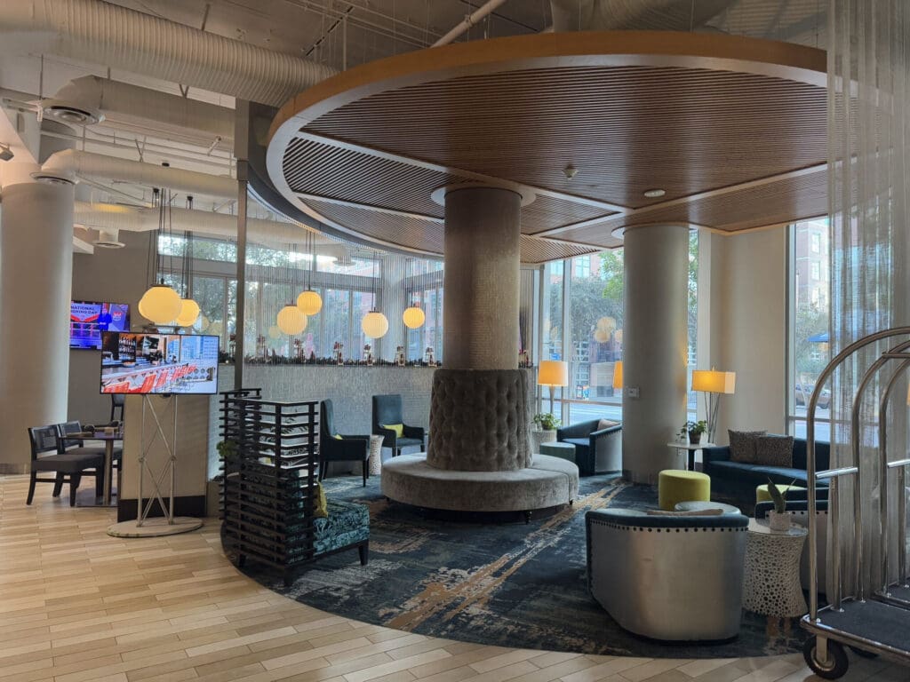 The image shows a modern hotel lobby with a stylish interior design. There is a circular seating area with a cushioned bench around a central column. The ceiling features a wooden circular design above the seating area. Several round pendant lights hang from the ceiling, providing warm lighting. The lobby has large windows allowing natural light to enter, and there are various chairs and small tables arranged around the space. A television is mounted on a stand, and a luggage cart is visible on the right side. The floor is covered with a patterned carpet and light-colored tiles.