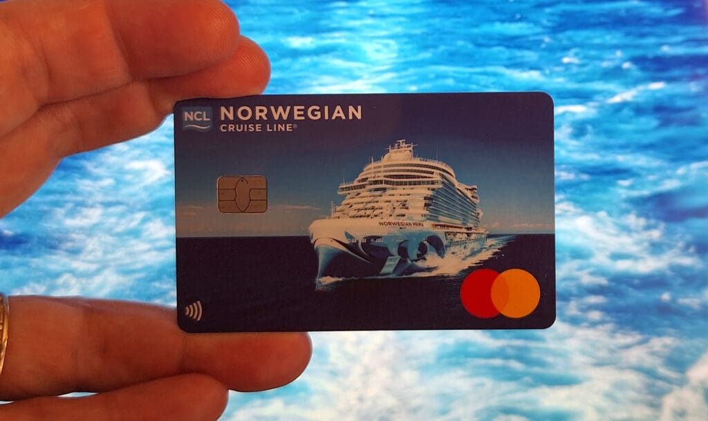 A hand is holding a credit card featuring an image of a cruise ship on the ocean. The card has the text "Norwegian Cruise Line" and "Norwegian Prima" on it, along with a chip and the Mastercard logo. The background shows a blurred view of the ocean.