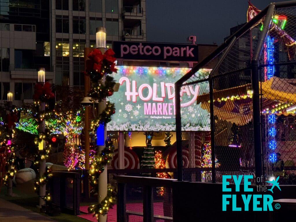 The image shows a festive holiday market scene at night, located at Petco Park. The area is decorated with colorful lights, wreaths, and garlands. A large sign reads "Holiday Market" with a candy cane design. There are illuminated trees and structures, creating a vibrant and cheerful atmosphere.