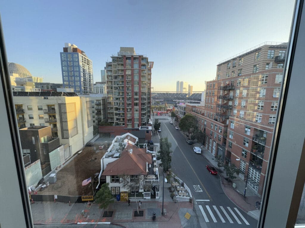 Hotel Indigo San Diego Gaslamp Quarter Studio Suite. A cityscape view from a window showing a street lined with modern buildings. In the foreground, there is a construction site with a fenced-off area and an American flag. The street has a few parked cars and a pedestrian crossing. In the background, a large stadium is visible, and the sky is clear with a hint of sunset light.