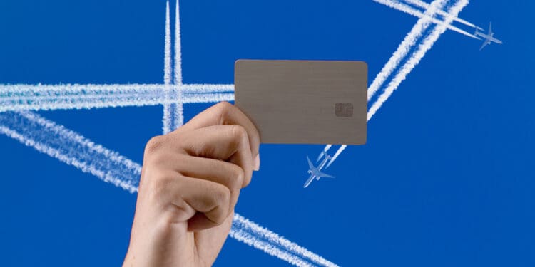 A hand is holding a credit card against a clear blue sky. In the background, several airplanes are flying, leaving white contrails that crisscross the sky.