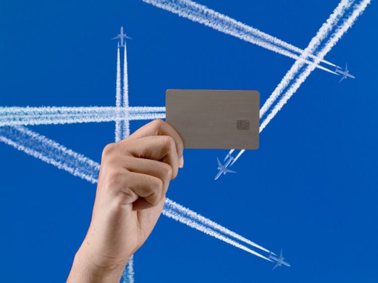 A hand is holding a credit card against a clear blue sky. In the background, several airplanes are flying, leaving white contrails that crisscross the sky.