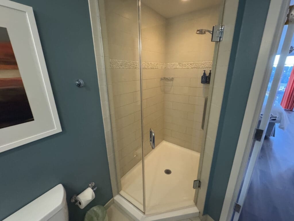Hotel Indigo San Diego Gaslamp Quarter Studio Suite. The image shows a bathroom with a glass-enclosed corner shower. The shower has beige tiles and a small shelf holding two bottles. The bathroom wall is painted blue, and there is a piece of framed artwork hanging on it. A toilet paper holder with a roll is visible, along with part of a toilet. The door to the bathroom is open, revealing a glimpse of a room with wooden flooring and red curtains.