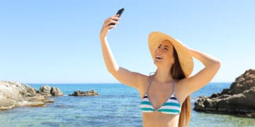 A woman wearing a striped bikini and a wide-brimmed straw hat is taking a selfie with her smartphone. She is standing by the ocean, smiling, with clear blue water and rocky formations in the background under a clear sky.