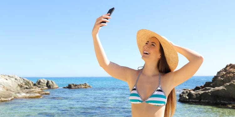 A woman wearing a striped bikini and a wide-brimmed straw hat is taking a selfie with her smartphone. She is standing by the ocean, smiling, with clear blue water and rocky formations in the background under a clear sky.