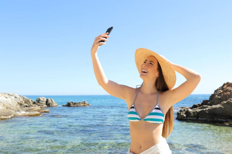 A woman wearing a striped bikini and a wide-brimmed straw hat is taking a selfie with her smartphone. She is standing by the ocean, smiling, with clear blue water and rocky formations in the background under a clear sky.