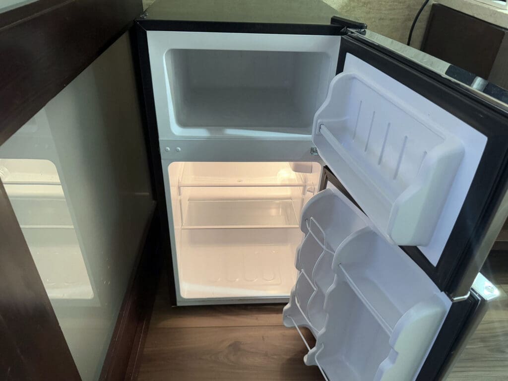 Hotel Indigo San Diego Gaslamp Quarter Studio Suite The image shows an open, empty mini refrigerator with a freezer compartment at the top. The fridge door has several shelves, and the interior is lit, revealing two main shelves inside. The refrigerator is positioned on a wooden floor next to a dark cabinet.
