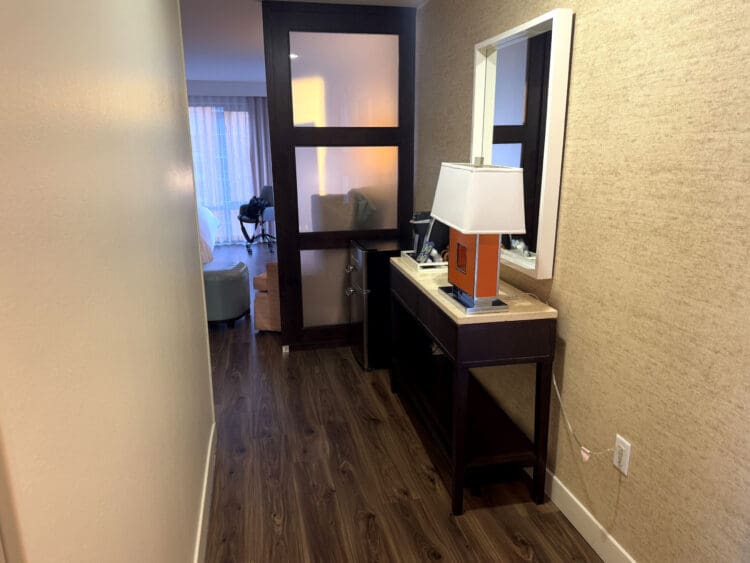 Hotel Indigo San Diego Gaslamp Quarter Studio Suite. A hotel room hallway with wooden flooring. On the right, there's a console table with a lamp and a mirror above it. The hallway leads to a room with a bed, a chair, and a window with curtains. A frosted glass partition partially separates the hallway from the room.