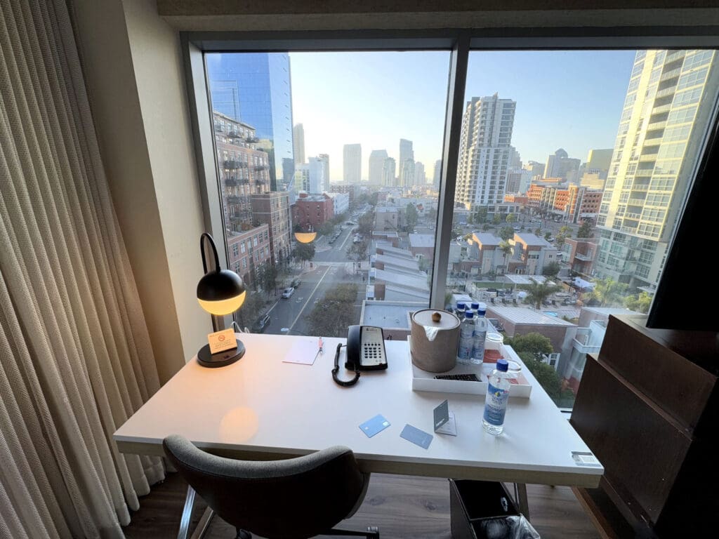 Hotel Indigo San Diego Gaslamp Quarter Studio Suite. A modern hotel room desk is positioned in front of a large window with a cityscape view. The desk features a black lamp, a telephone, a notepad with a pen, and several water bottles. An ice bucket and a few cards are also on the desk. The window offers a view of tall buildings and a street below, with a clear sky in the background. A chair is placed in front of the desk, and curtains are partially visible on the left side.