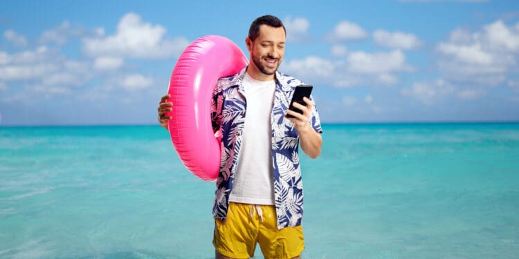 A man standing in shallow ocean water, holding a pink inflatable ring and looking at his smartphone. He is wearing a blue and white floral shirt and yellow shorts. The sky is clear with a few clouds, and the water is a bright turquoise.