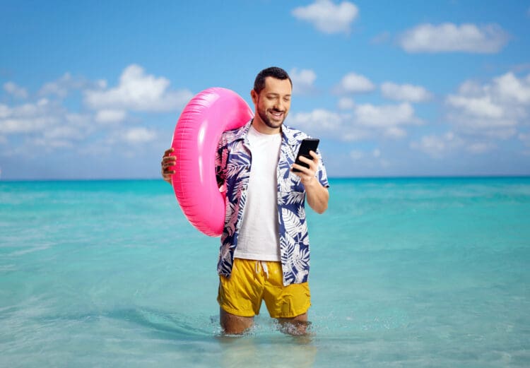 A man standing in shallow ocean water, holding a pink inflatable ring and looking at his smartphone. He is wearing a blue and white floral shirt and yellow shorts. The sky is clear with a few clouds, and the water is a bright turquoise.
