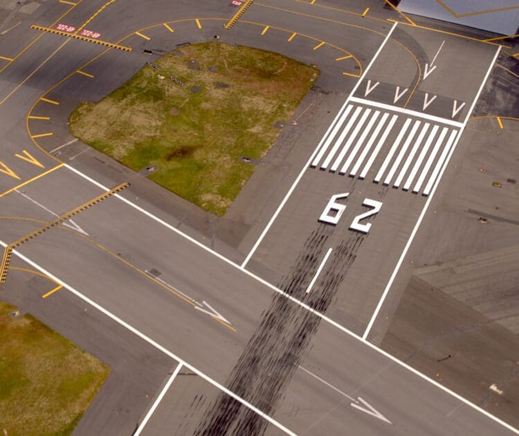 Aerial view of an airport runway with the number "62" painted on it. The runway is marked with white lines and arrows, and there are visible tire skid marks. Surrounding the runway are taxiways with yellow markings and a grassy area.