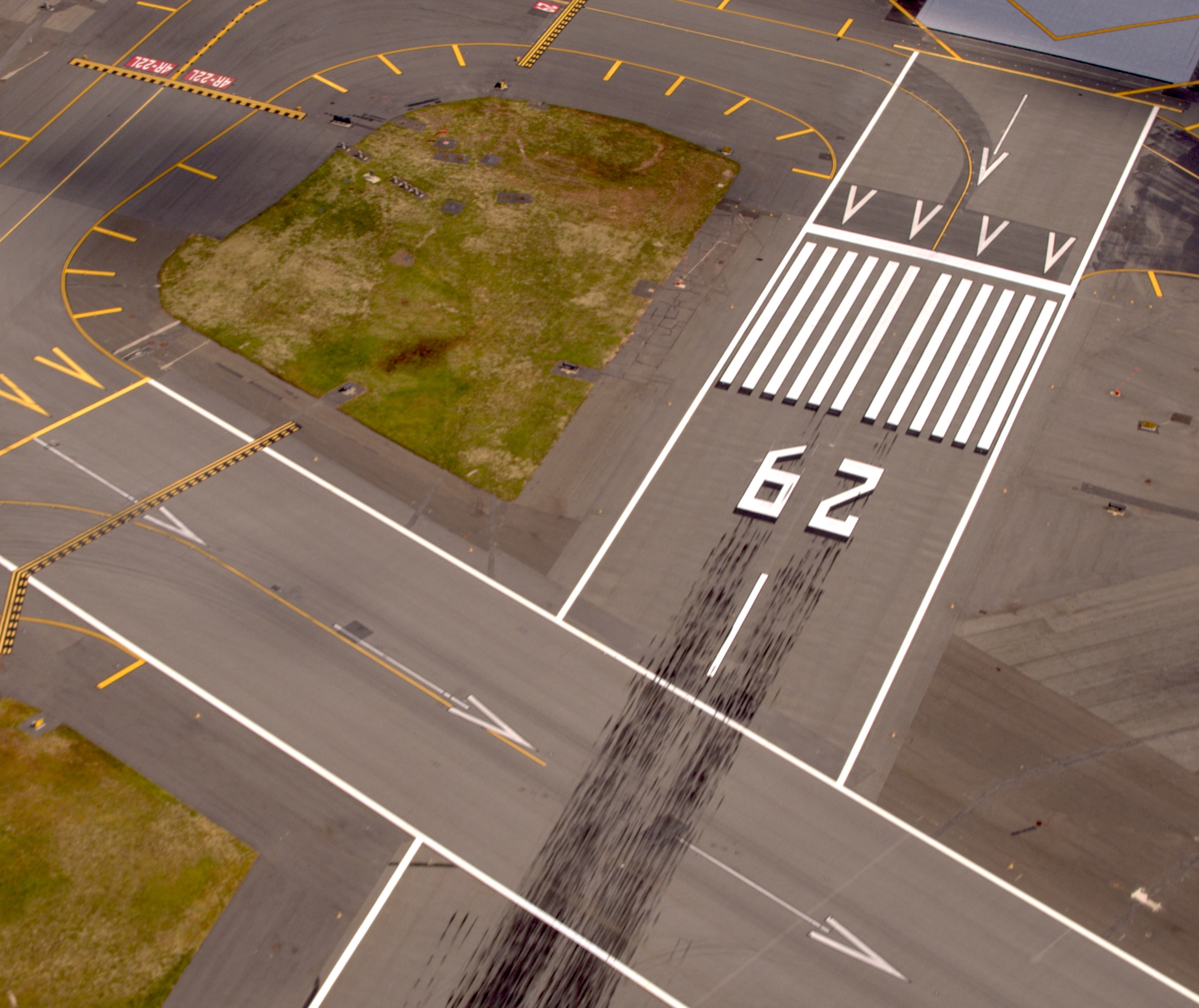 Aerial view of an airport runway with the number "62" painted on it. The runway is marked with white lines and arrows, and there are visible tire skid marks. Surrounding the runway are taxiways with yellow markings and a grassy area.