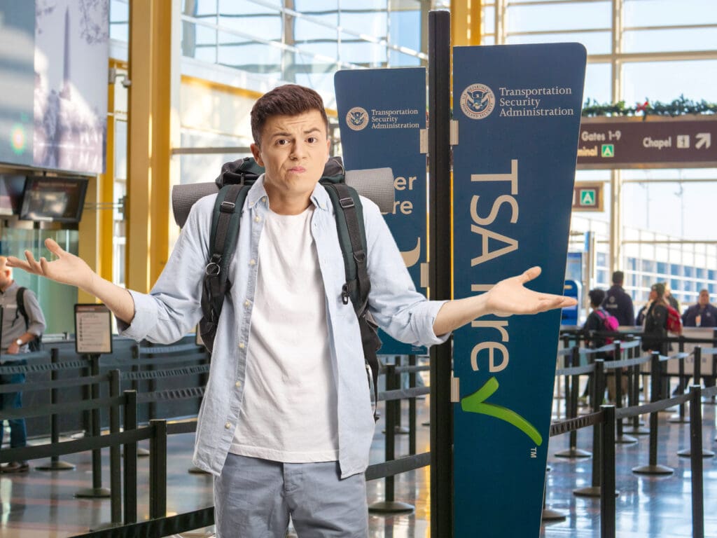 A person with a backpack is standing in an airport near a TSA PreCheck sign. They have a confused expression and are shrugging their shoulders. The background shows airport signage and people in line.