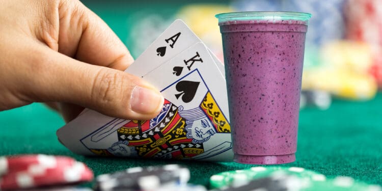 A hand holding a royal flush in spades (Ace, King, Queen) is shown next to a purple smoothie in a plastic cup. The background features a green poker table with scattered poker chips.