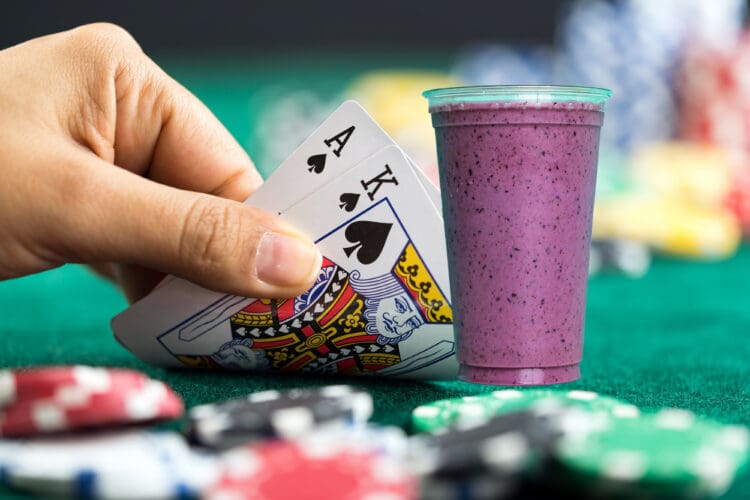 A hand holding a royal flush in spades (Ace, King, Queen) is shown next to a purple smoothie in a plastic cup. The background features a green poker table with scattered poker chips.