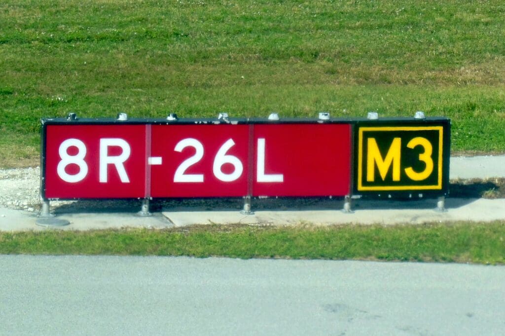 The image shows an airport taxiway sign. The sign has a red background with white text displaying "8R-26L" and a black background with yellow text displaying "M3." The sign is situated on a grassy area next to a paved surface.