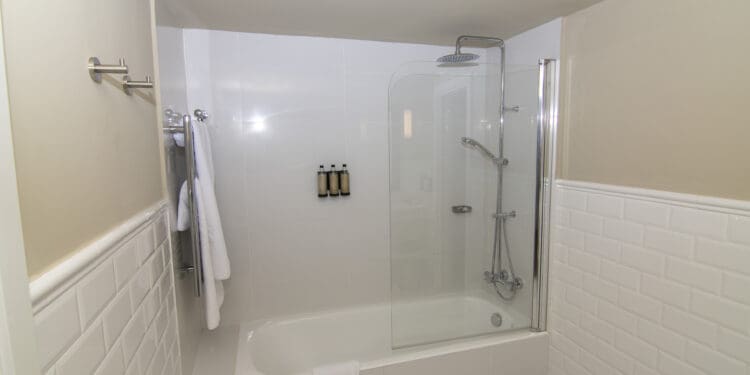 The image shows a modern bathroom with a white bathtub and a glass shower enclosure. There is a wall-mounted showerhead and a handheld shower attachment. Three bottles of toiletries are mounted on the wall inside the shower. A white towel is draped over the edge of the bathtub, and another towel is hanging on a rack outside the shower. The walls are tiled with white subway tiles.