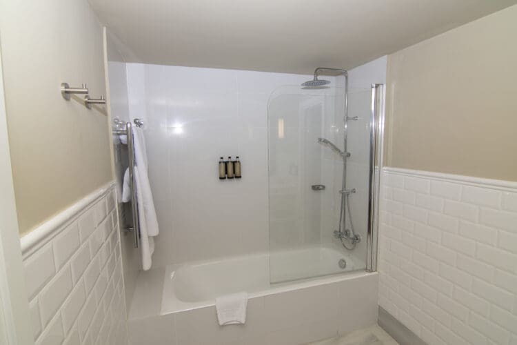 The image shows a modern bathroom with a white bathtub and a glass shower enclosure. There is a wall-mounted showerhead and a handheld shower attachment. Three bottles of toiletries are mounted on the wall inside the shower. A white towel is draped over the edge of the bathtub, and another towel is hanging on a rack outside the shower. The walls are tiled with white subway tiles.