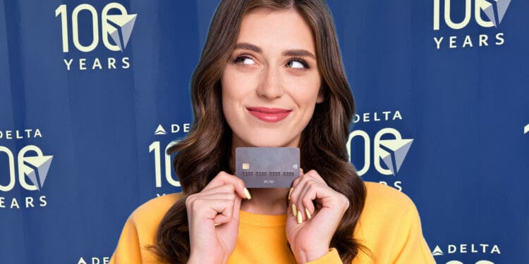 A woman in a yellow sweater is holding a credit card and smiling. She is standing in front of a blue backdrop with the text "DELTA 100 YEARS" repeated multiple times.