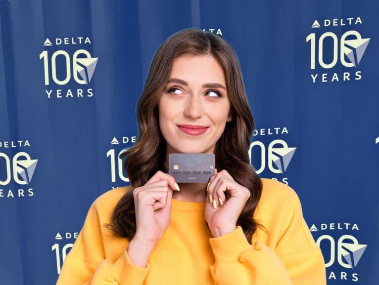 A woman in a yellow sweater is holding a credit card and smiling. She is standing in front of a blue backdrop with the text "DELTA 100 YEARS" repeated multiple times.