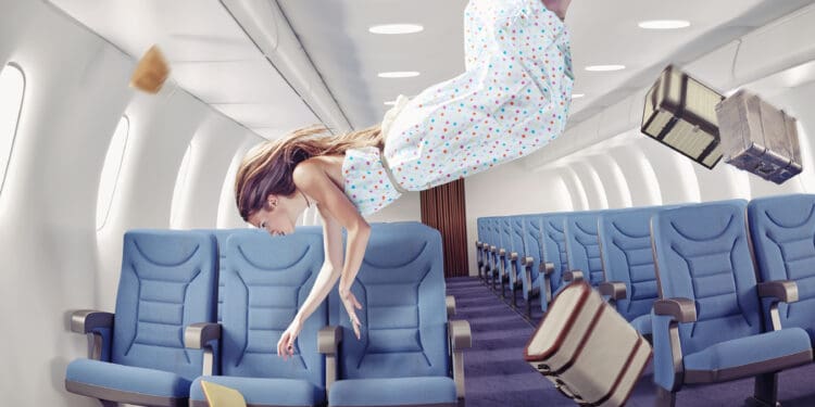 A woman in a polka dot dress is floating in the aisle of an airplane cabin, as if experiencing zero gravity. Blue airplane seats line both sides of the aisle. Several objects, including suitcases and a book, are also floating in the air. The scene gives a surreal impression of weightlessness inside the aircraft.