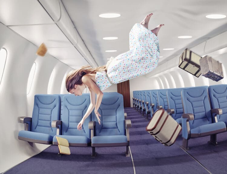 A woman in a polka dot dress is floating in the aisle of an airplane cabin, as if experiencing zero gravity. Blue airplane seats line both sides of the aisle. Several objects, including suitcases and a book, are also floating in the air. The scene gives a surreal impression of weightlessness inside the aircraft.