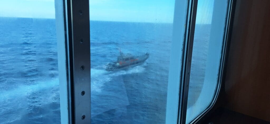 The image shows a view through a window of a ship, looking out onto the ocean. A small pilot boat is visible on the water, creating a wake as it moves. The sea is calm, and the sky is clear.