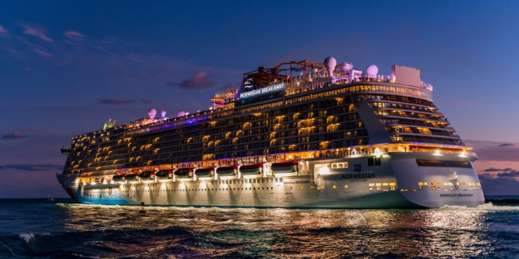A large cruise ship, the Norwegian Breakaway, is illuminated against a twilight sky. The ship is brightly lit with numerous lights along its decks, reflecting on the water below. The sky is a deep blue with a few clouds visible.