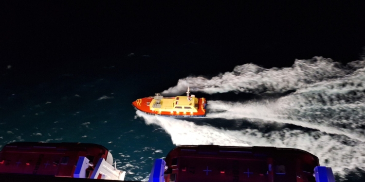 A small, brightly lit pilot boat with a red and yellow color scheme is moving through dark waters at night, creating a trail of white waves behind it. The image is taken from above, possibly from a larger vessel, parts of which are visible at the bottom of the image.