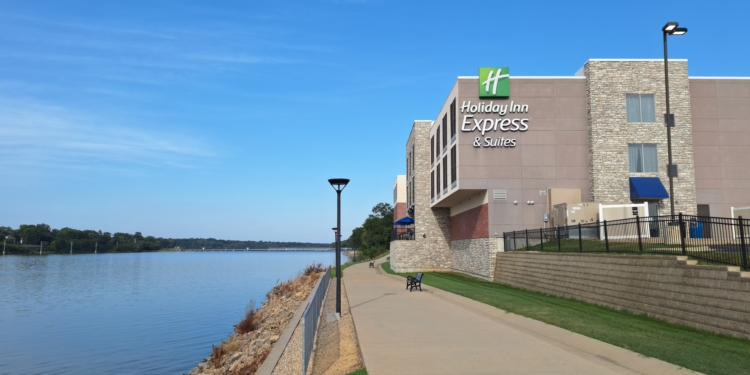 The image shows a Holiday Inn Express & Suites building located next to a river. The hotel has a modern design with stone and brick elements. A paved walkway runs alongside the river, bordered by a metal fence and grassy areas. The sky is clear and blue, suggesting a sunny day.