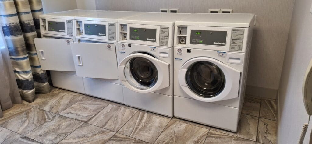 The image shows a row of three white front-loading washing machines in a laundry room. Two of the machines have their doors open, revealing the drum inside. The machines are placed on a tiled floor, and there are patterned curtains on the left side of the image.