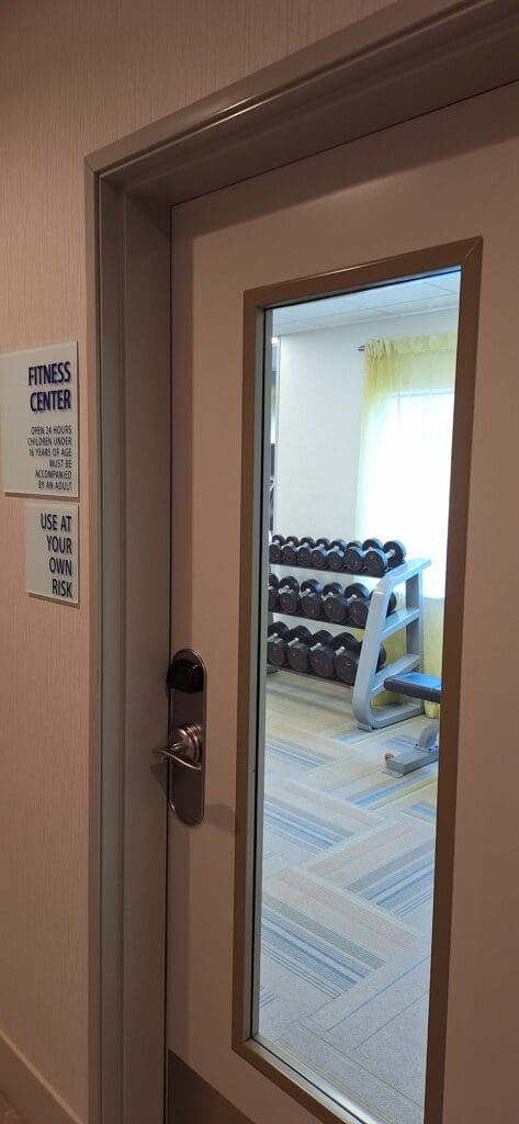 The image shows a door with a window, leading to a fitness center. Through the window, a rack of dumbbells is visible inside the room. There is a sign on the door that reads "Fitness Center" and mentions it is open 24 hours, with a note that children under 16 must be accompanied by an adult. Another sign says "Use at your own risk."