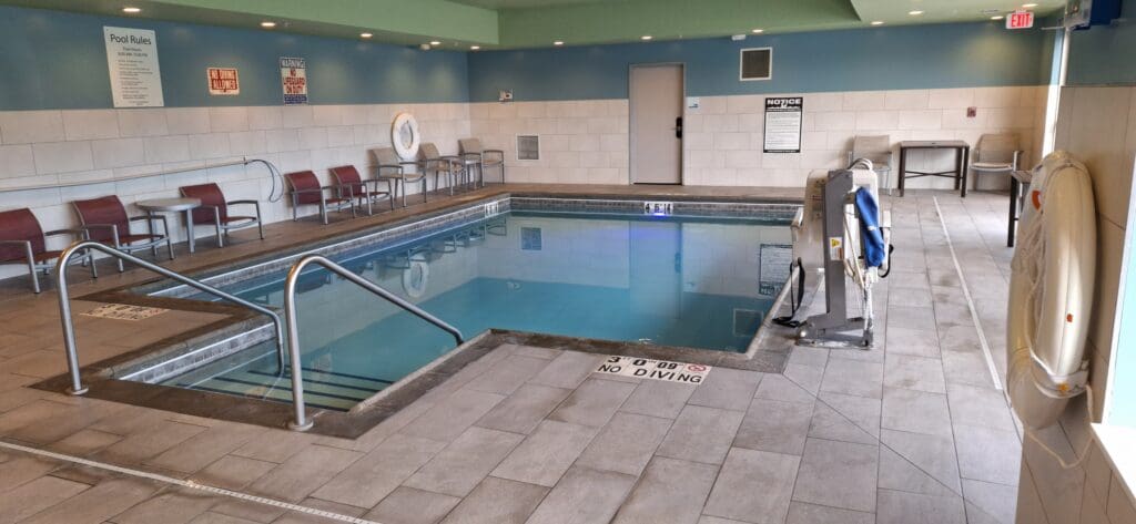 The image shows an indoor swimming pool area with a small rectangular pool. The pool is surrounded by a tiled floor and has metal handrails for entry. There are several chairs and tables along the wall, and a pool lift is visible on the right side. Signs on the wall include pool rules and a warning about no lifeguard on duty. A life preserver is mounted on the wall, and there is an exit sign above a door in the background.