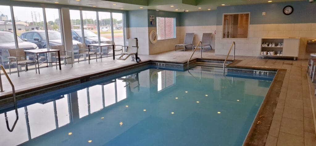 The image shows an indoor swimming pool area with clear water and metal handrails leading into the pool. There are several lounge chairs and tables along the side, with large windows providing a view of parked cars and buildings outside. The walls are tiled, and there is a clock and a shelf with towels on the right side. The pool depth is marked as 3 feet.